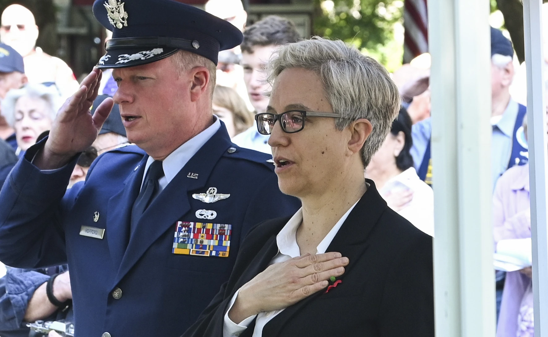 Tina Kotek stands with her hand over her chest during a public ceremony beside a saluting U.S. military officer, with attendees gathered in the background.
